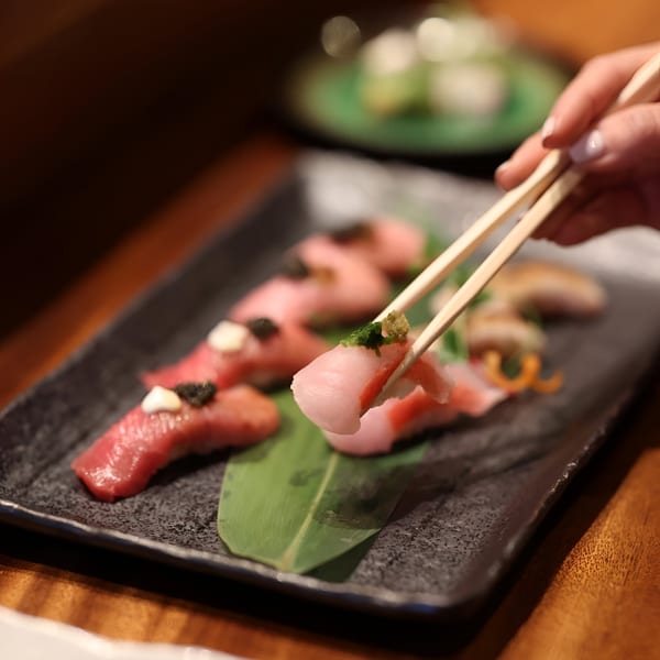 Chopsticks picking up a piece of MF Sushi nigiri from a black plate with other sushi.